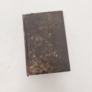 May include: A small, antique book with a dark brown, textured cover. The cover shows signs of wear and age, with faded areas and a slightly distressed appearance. The book is rectangular and appears to be closed.