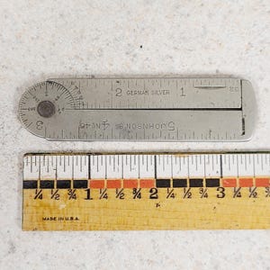 May include: A vintage metal protractor and ruler set. The protractor is marked with degree measurements and the text "GERMAN SILVER". A wooden ruler with inch markings is below the protractor. The ruler is marked "MADE IN U.S.A."