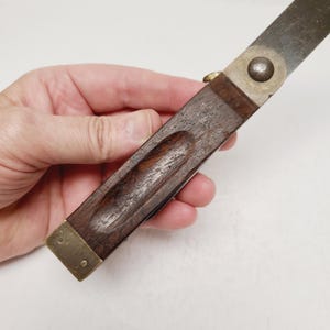 May include: A woodworking tool featuring a dark brown wooden handle with a recessed grip area, accented by a brass end cap and a brass pivot joint that connects to a flat metal blade extending from the top of the handle structure.