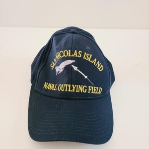 May include: Navy blue baseball cap with embroidered text and graphics. The text reads "SAN NICOLAS ISLAND NAVAL OUTLYING FIELD" in yellow. The cap features a curved brim and a six-panel design.