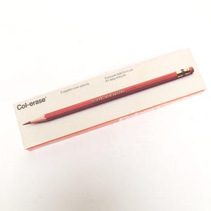 May include: A rectangular off-white box featuring a red erasable color pencil with a metal ferrule and eraser. The text on the box reads Col-erase, Erasable color pencils, and Exclusive lead formula for easy erasure in black lettering.