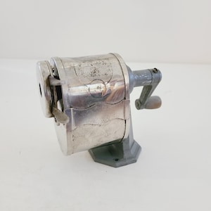 Puede incluir: Un sacapuntas vintage de plata con una manivela y una base de metal. El sacapuntas tiene una etiqueta que dice "Victor, Chicago, Ill. U.S.A."