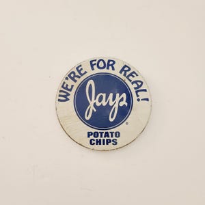 May include: A round, vintage Jay's Potato Chips sign with a white background and blue lettering. The text reads "WE'RE FOR REAL!" around the edge, with "Jay's" in a central blue circle, and "POTATO CHIPS" below.