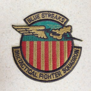 May include: Embroidered patch featuring the text "BLUE STREAKS" above a design of wings and a stylized cloud. The patch has a red and gold striped shield with the text "91st TACTICAL FIGHTER SQUADRON" around the bottom edge. The patch is in shades of green, gold, red, and black.