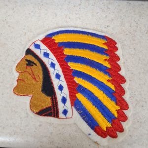 May include: An embroidered fabric patch featuring the profile of a person with tan skin and black hair, wearing a traditional headdress with alternating yellow and blue feathers, a red border, and a white headband with blue diamond patterns.