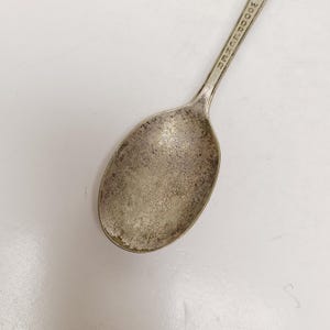 May include: Close-up of a vintage silver spoon with the word "WOODUCKER" engraved on the handle. The spoon has a worn, tarnished appearance, suggesting age and use. The spoon's bowl is oval-shaped.