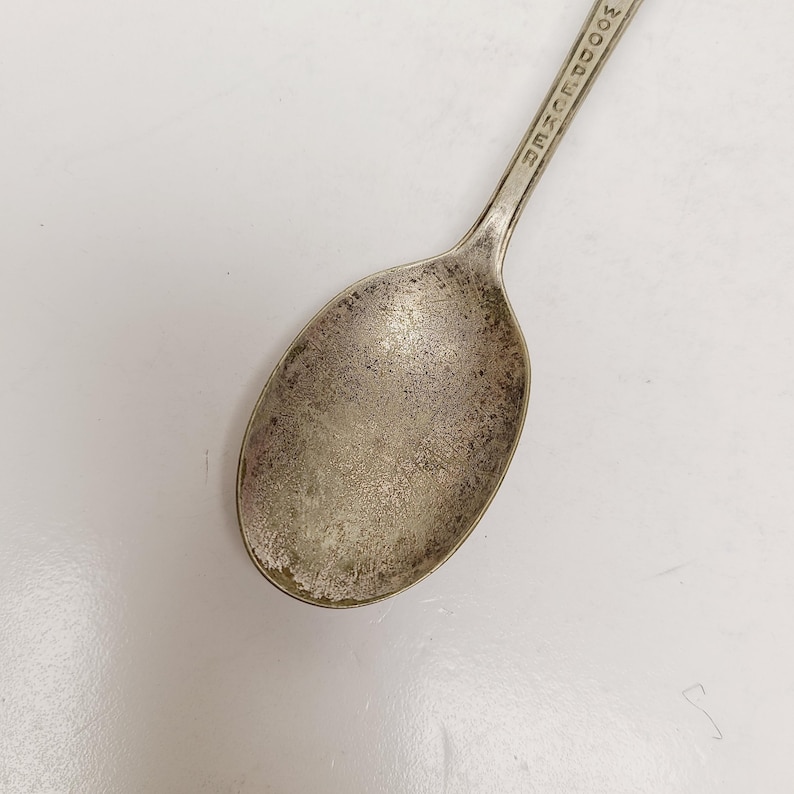 May include: Close-up of a vintage silver spoon with the word "WOODUCKER" engraved on the handle. The spoon has a worn, tarnished appearance, suggesting age and use. The spoon's bowl is oval-shaped.