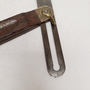 May include: A vintage sliding T-bevel tool featuring a dark brown wooden handle and a metallic blade. The two components are connected by a brass locking nut and a pivot mechanism, allowing the blade to be adjusted and secured at various angles.