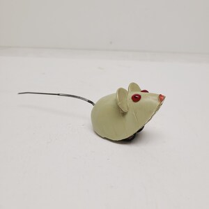 May include: A vintage-style wind-up toy mouse in a pale cream color with bright red painted eyes and a small red nose. The mouse features two rounded ears and a long, thin, flexible metal spring tail extending from the back of its rounded body.