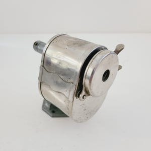 Puede incluir: Un sacapuntas vintage de plata con manivela y base de metal. El sacapuntas tiene una abertura redonda para l&aacute;pices y una cuchilla de metal.
