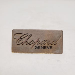 May include: A rectangular, brown wooden plaque with the brand name "Chopard" in a cursive font. Below the name, the word "GENÈVE" is printed in a smaller, black font. The plaque has a distressed, aged appearance.