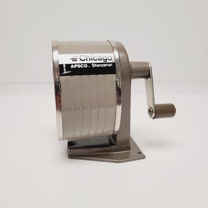 May include: A vintage Chicago APSCO pencil sharpener with a beige cylindrical body and a dark gray textured metal base. The device features a side-mounted hand crank with a brown handle and a black label with white text reading Chicago APSCO Sharpener.