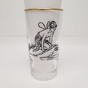 May include: A clear glass tumbler featuring a gold-toned rim and a black line-art illustration of a winged figure kneeling on a rock. The figure has short hair and is positioned near water with small plants on either side of the central rock formation.