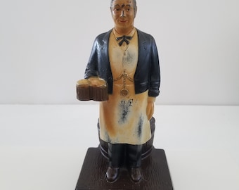 May include: A vintage figurine of a man in a black suit and apron holding a beer stein. The figurine is standing on a base with the text "Schmidt's Beer of Philadelphia Ale".