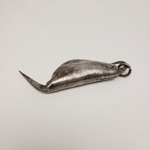 May include: A vintage, weathered metal tool with a curved, pointed blade and a rounded body. The tool has a small ring at the end and shows signs of age and use. The metal has a dull, gray finish.