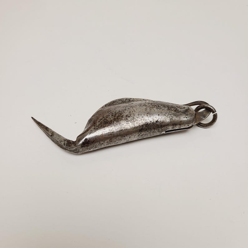 May include: A vintage, weathered metal tool with a curved, pointed blade and a rounded body. The tool has a small ring at the end and shows signs of age and use. The metal has a dull, gray finish.