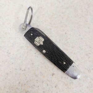 May include: A black pocket knife with a silver blade and a loop for attaching to a keychain. The handle has a textured pattern and a silver emblem with the letters "GS" inside a flower shape. The knife is closed and resting on a light-colored surface.