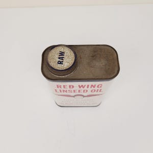 May include: A vintage tin can of Red-Wing Linseed Oil. The can is white with red lettering and a red wing logo. The can has a rusty top and a blue and white lid with the word "RAW" printed on it.