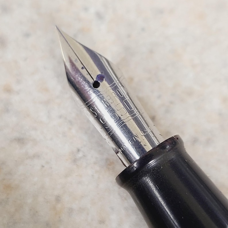 May include: Close-up of a silver fountain pen nib with a black barrel. The nib has a fine point and is marked with text. The pen is designed for writing and calligraphy.