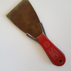 May include: A vintage Red Devil scraper with a worn, rectangular metal blade and a red wooden handle. The handle has the brand name and product information printed on it. The tool is designed for scraping and is likely used for home improvement or construction tasks.