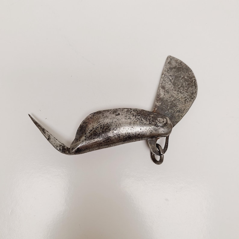May include: An antique, metal tool with a curved blade and a pointed end. The tool has a hinged, semi-circular blade and a small ring attached to the body. The metal has a worn, aged appearance.