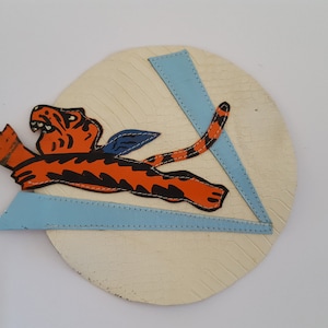 May include: A round, white, wooden plaque with a blue and white triangle design. A cartoon orange and black tiger with blue wings is leaping across the triangle.