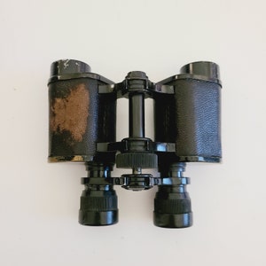 May include: Black binoculars with a leather covering on the barrels. The binoculars have a center focus knob and a strap loop.