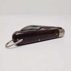 May include: A vintage pocket knife with a dark brown handle and a partially open blade. The knife has a silver-colored metal end and a metal loop for attaching to a keychain. The blade shows signs of wear.