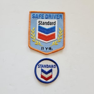 May include: Two embroidered patches featuring the Standard Oil logo. The larger patch is blue with yellow trim and reads "SAFE DRIVER Standard 11 YR.". The smaller patch is blue with a white chevron and the word "STANDARD" in white.