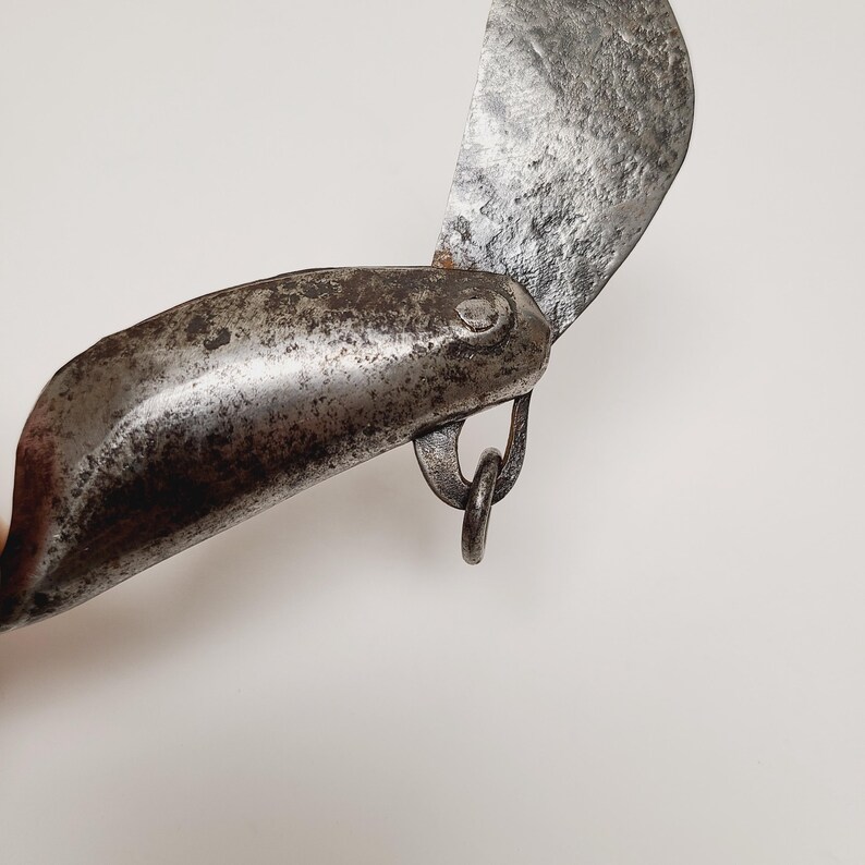 May include: Close-up of a vintage, weathered metal knife with a curved blade. The knife features a dark, aged appearance with visible wear and tear, suggesting its antique nature. A small metal ring is attached to the handle.