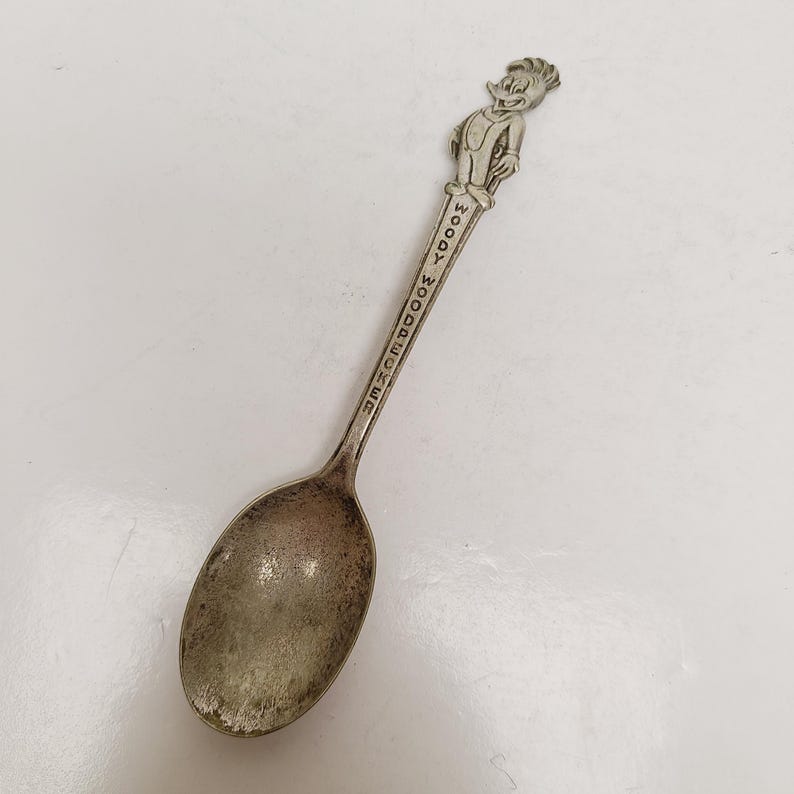 May include: A vintage silver spoon featuring a detailed figure of Woody Woodpecker at the handle's top. The spoon's handle is inscribed with "WOODY WOODPECKER". The spoon has a classic oval bowl shape and shows signs of age.
