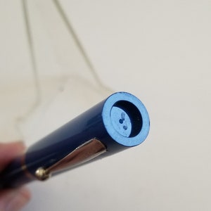 May include: A close-up of a navy blue fountain pen with a gold-colored clip and trim. The pen has a cylindrical shape and a dark blue end with three small holes. The pen is held at an angle.