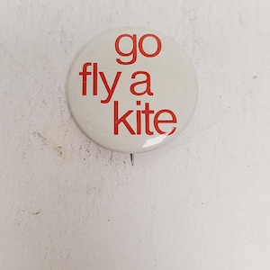 May include: A round, off-white button with the red text "go fly a kite". The button has a pin on the back. The background is a textured, off-white surface.