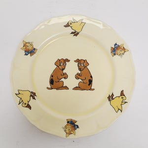 May include: A cream-colored ceramic plate with a scalloped edge. The plate features cartoon illustrations of two brown dogs, yellow ducks, and orange cats with blue bows. The plate is a charming piece of tableware.