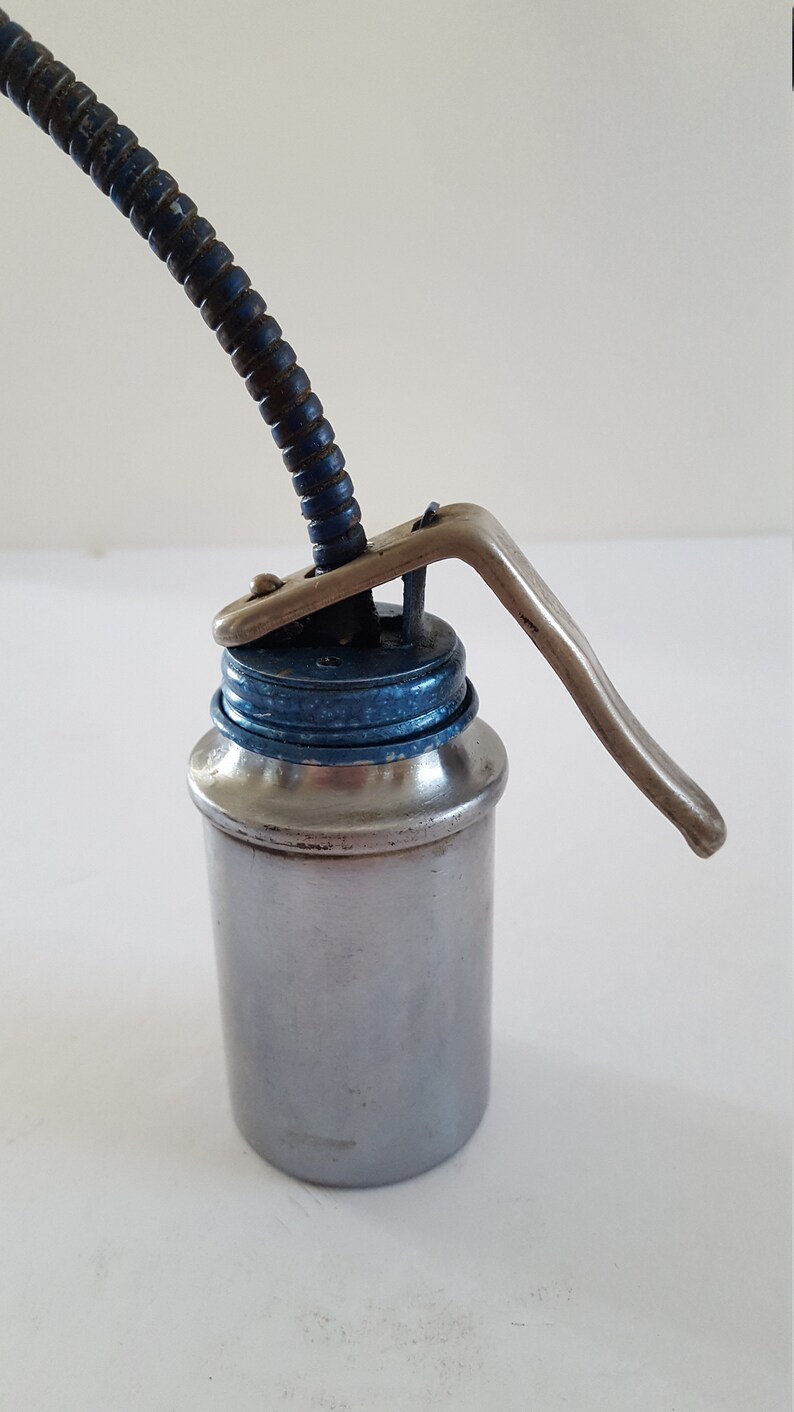 Puede incluir: Una lata de aceite plateada con una tapa azul y un pico largo y flexible. La lata tiene una palanca en el lateral para bombear aceite.