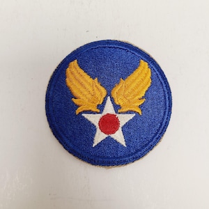 May include: A circular patch with a blue background featuring a white star with a red center and gold wings. The patch is embroidered with a brown border.