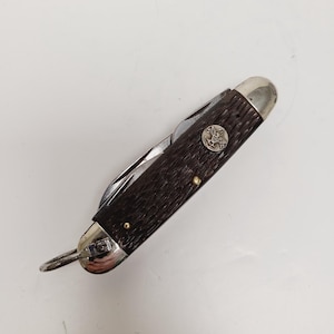 May include: A vintage pocket knife with a dark brown handle and a silver-colored blade. The knife features a metal emblem with an eagle design and a small ring for attaching to a keychain. The knife is closed, with the blade partially visible.
