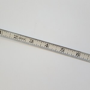 Puede incluir: Una cinta m&eacute;trica Lufkin blanca, marcada en pulgadas, se encuentra en diagonal sobre una superficie blanca. La cinta muestra medidas de 2.5 cm a 20.3 cm. Fabricada en EE. UU.