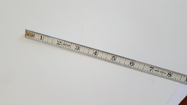Puede incluir: Una cinta m&eacute;trica Lufkin blanca, marcada en pulgadas, se encuentra en diagonal sobre una superficie blanca. La cinta muestra medidas de 2.5 cm a 20.3 cm. Fabricada en EE. UU.