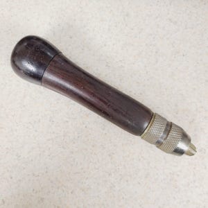 May include: A vintage hand drill with a dark wooden handle and a metal chuck. The handle is a dark brown color, and the metal chuck has a textured grip. The drill is designed for manual operation.