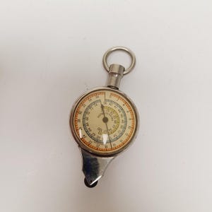 May include: A vintage silver-toned map measurer with a circular dial. The dial features measurements in inches and a rotating needle. The device has a small metal loop at the top and a small wheel at the bottom.
