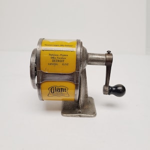 May include: A vintage metal pencil sharpener with a yellow cylindrical body featuring black text that reads Giant, Gregory Mayer & Thom Co., Stationers, Printers, Office Furniture, Detroit, Lansing, Flint, and America's Largest Office Outfitters.