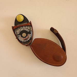 May include: A vintage light meter with a brown leather case. The meter has a black face with white and red markings, a yellow dome, and a leather strap. The case is open, revealing the meter's face and dials.