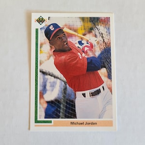 Puede incluir: Tarjeta de béisbol con Michael Jordan con camiseta roja y gorra azul, en pleno swing con un bate. La tarjeta tiene un borde blanco con detalles verdes y naranjas. El texto "Michael Jordan" está en la parte inferior.