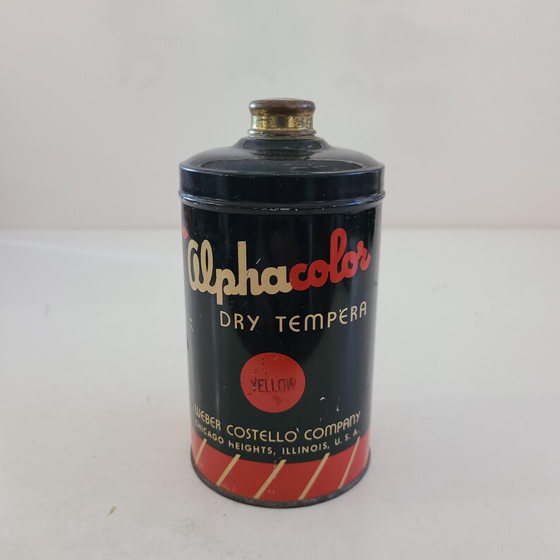 May include: A vintage tin of Alphacolor Dry Tempera paint in yellow. The tin is black with red and white stripes and the text "Alphacolor Dry Tempera" and "Weber Costello Company Chicago Heights, Illinois, U.S.A."