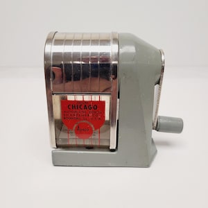May include: A vintage grey metal pencil sharpener with a chrome-plated top and a clear plastic shavings receptacle. A red label on the front reads: Deluxe Chicago Automatic Pencil Sharpener Co., Rockford, Ill., U.S.A. and features the Apsco logo.
