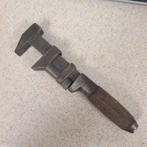 May include: A vintage adjustable wrench featuring a dark, weathered metal head and a cylindrical wooden handle. The tool includes a knurled adjustment nut located between the metal jaw mechanism and the handle, showing signs of age and wear.