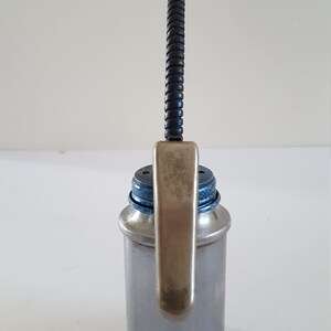 Puede incluir: Una lata de aceite plateada con un pico flexible negro y una palanca plateada. La lata tiene una tapa azul.