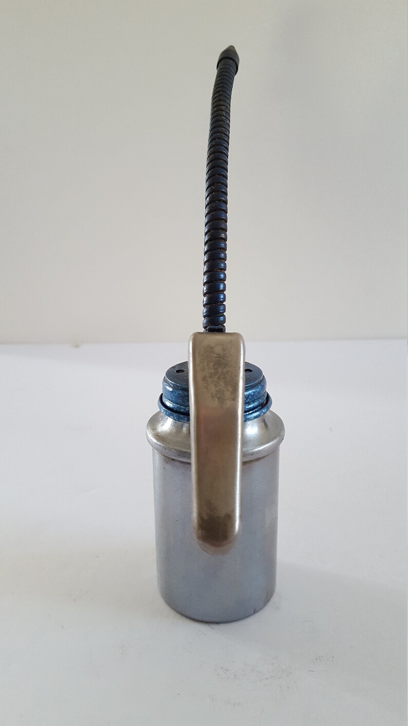 Puede incluir: Una lata de aceite plateada con un pico flexible negro y una palanca plateada. La lata tiene una tapa azul.