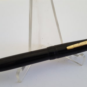 May include: A black fountain pen with gold-colored accents and the text "BOOK-FOX" on the clip. The pen is displayed on a clear acrylic stand against a white background. The pen's design is sleek and classic.
